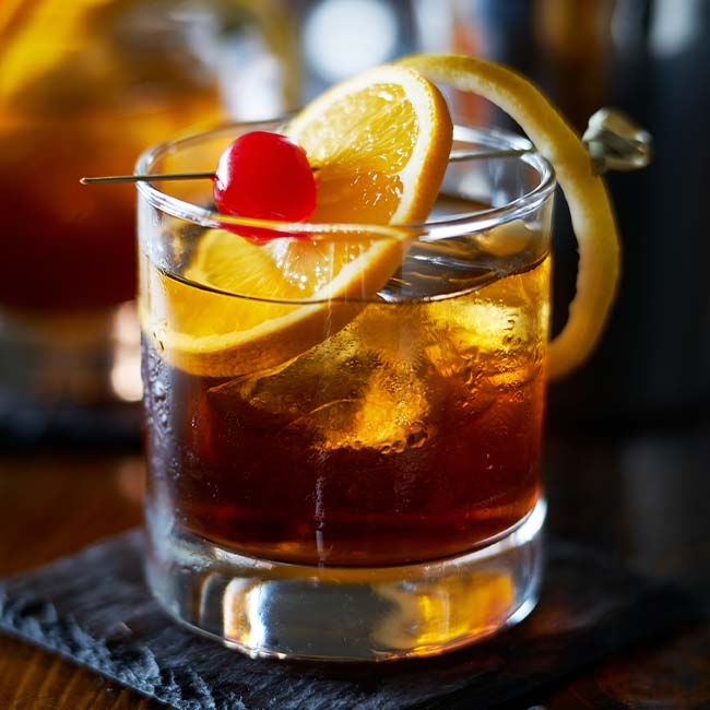 Glass of dark cocktail with ice, orange slice, and cherry at a Green Bay family restaurant.