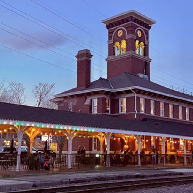 A brick train station, The Depot, with a clock tower and outdoor bar lit by string lights at dusk.